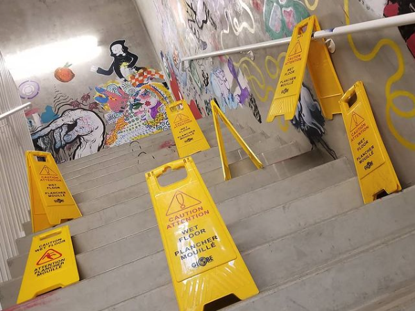 Seven yellow plastic “wet floor” hazard signs are placed haphazardly, blocking a set of stairs in a concrete hallway. The walls are painted with colourful murals.