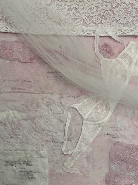 A canvas covered in pink fabric, white lace, and tissue paper with a parchment paper bra draped across the corner.