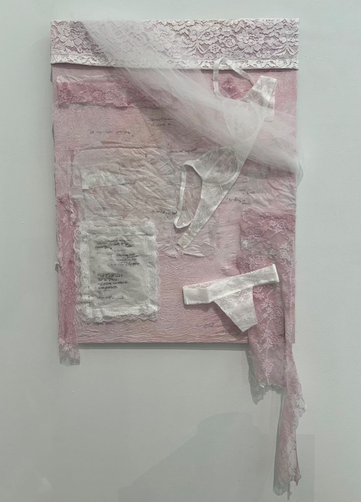 An image of bedroom, A canvas covered in pink fabric, white lace, and tissue paper with a parchment paper underwear set and a pillow, with a poem written on it.