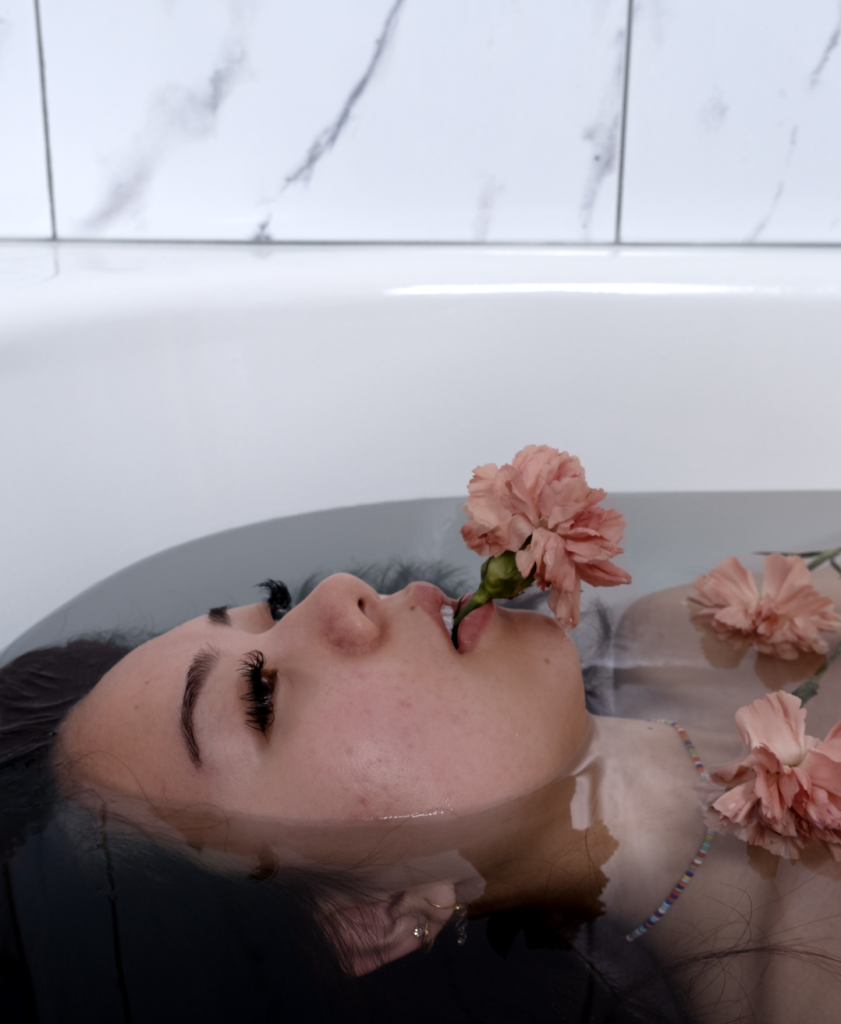 a woman's face half emerged in a bathtub with an orange flower coming out of her mouth