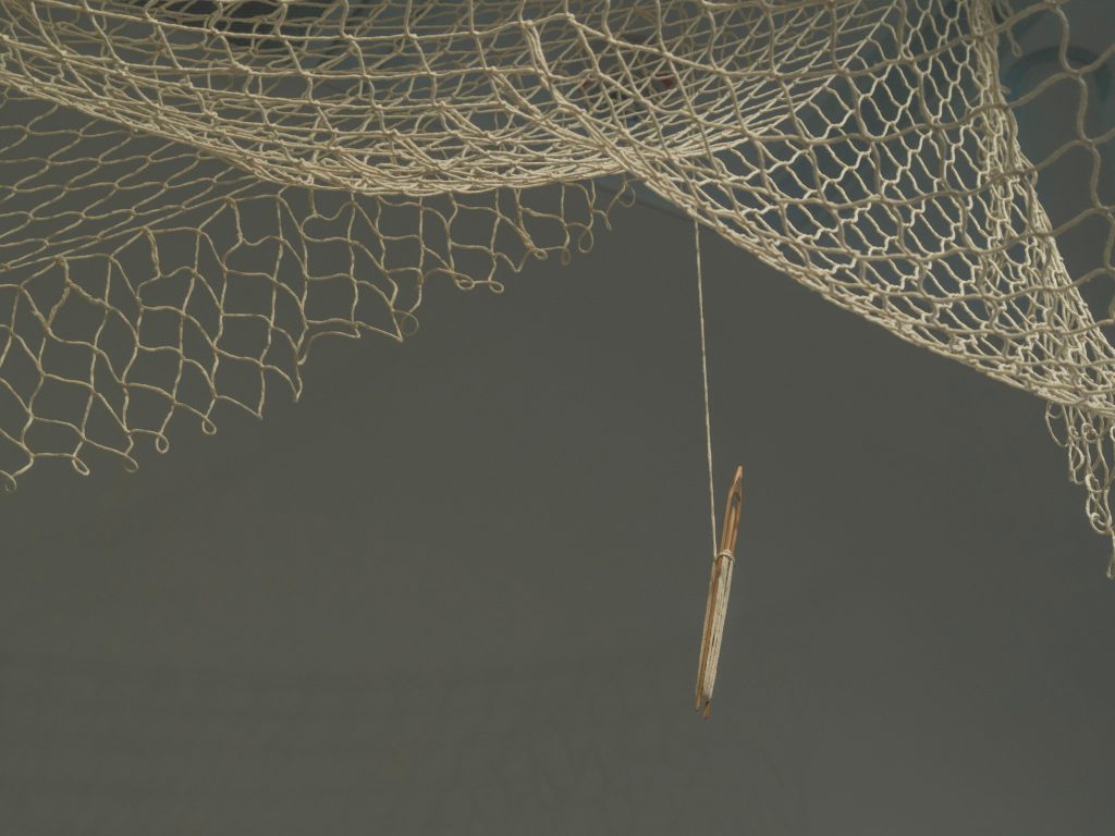Image of "Temporary Suspension" (2024), a sculptural artwork of cotton twin netting and netting shuttles suspended from the ceiling.