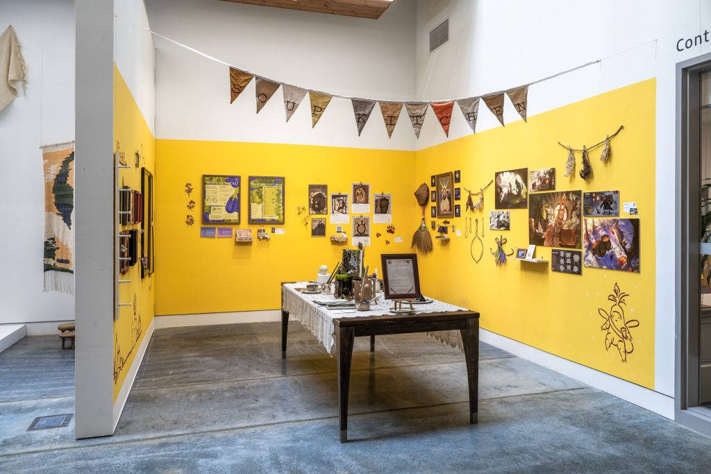 Gallery installation view showing a space enclosed by three walls painted bright yellow. Mounted on the walls are a wide assortment of pictures and artifacts, as well as a line drawing of a winged anthropomorphic character. In the centre of the enclosure is a wood table with white tablecloth displaying an assortment of artifacts and a framed document. Bunting flags drawn across the opening of the enclosure spell out "Soup Kitchen"
