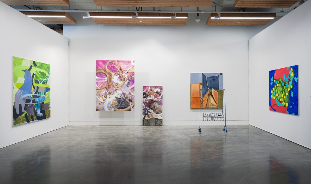Gallery installation view. There is a set of five abstract paintings on the white wall. On the left is a painting of varying shades of blue and green. To the left of the center are two paintings in groups of pink, white, brown, and silver. One of the smaller ones is supported by two squares. To the right of the center is a painting consisting of green blue and orange, which has a thin blue shelf placed in front of it. To the far right is a painting composed of highly saturated colors, there is a blue background dotted with large swaths of red and shades of green, and the shape of the subject looks like an apple.