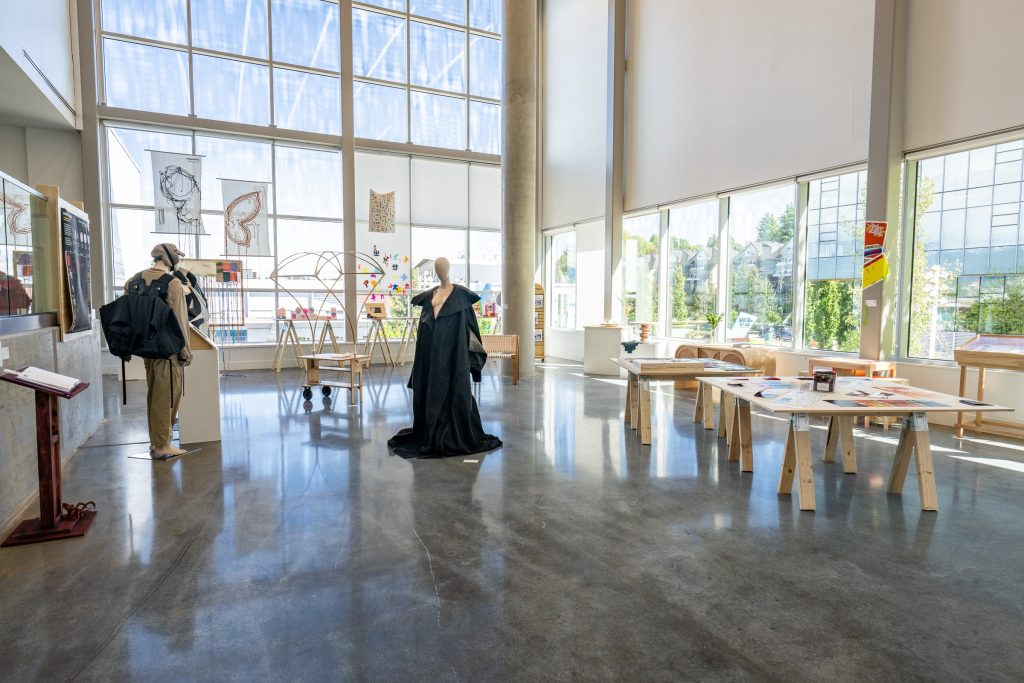 Gallery installation view. In the foreground are two mannequins, one wearing a large black backpack and tan clothing, and the other a flowing black robe. Also on display throughout the room are wooden tables and chairs. In the background are a variety of display tables showing small objects, and against the tall windows are weavings with trailing threads and decals of colourful letters. On the right, large wooden tables have printed materials spread out for perusal.