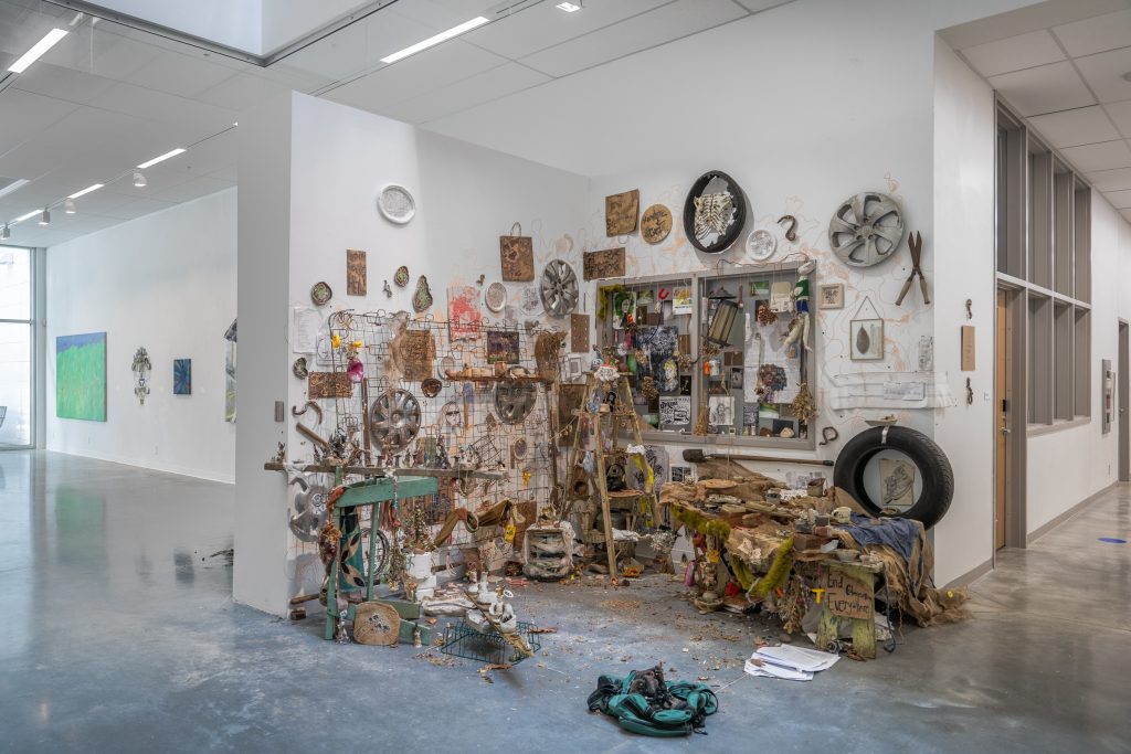 Installation view of a corner space with a chaotic and highly varied assembly of physical items, including hubcaps, a mattress frame, a tire, a ladder, and many others. The artifacts appear in various states of decay or weathering.