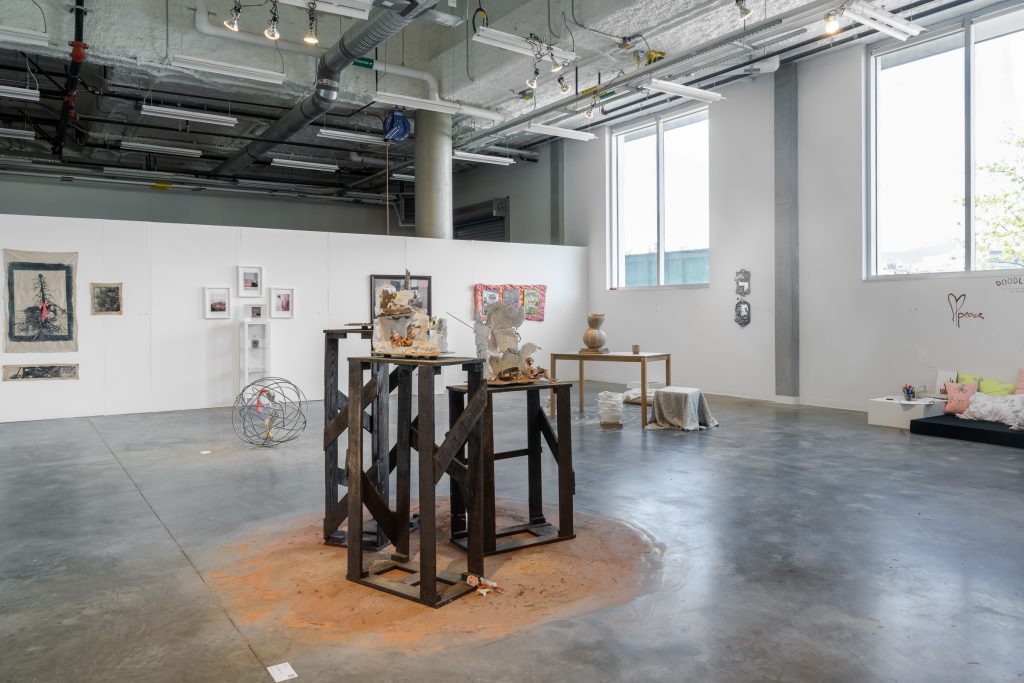 Gallery installation view. In the foreground are three high brown and black display shelves holding three different ceramic pieces. There is light brown sandy soil under and around the shelves. To the left rear of the shelves is a spherical device made of steel wire placed on the floor. On the right rear side is a wooden table next to a gray cloth placed on top of a ceramic piece. The final wall displays four 2D pieces. They are presented in different textures.