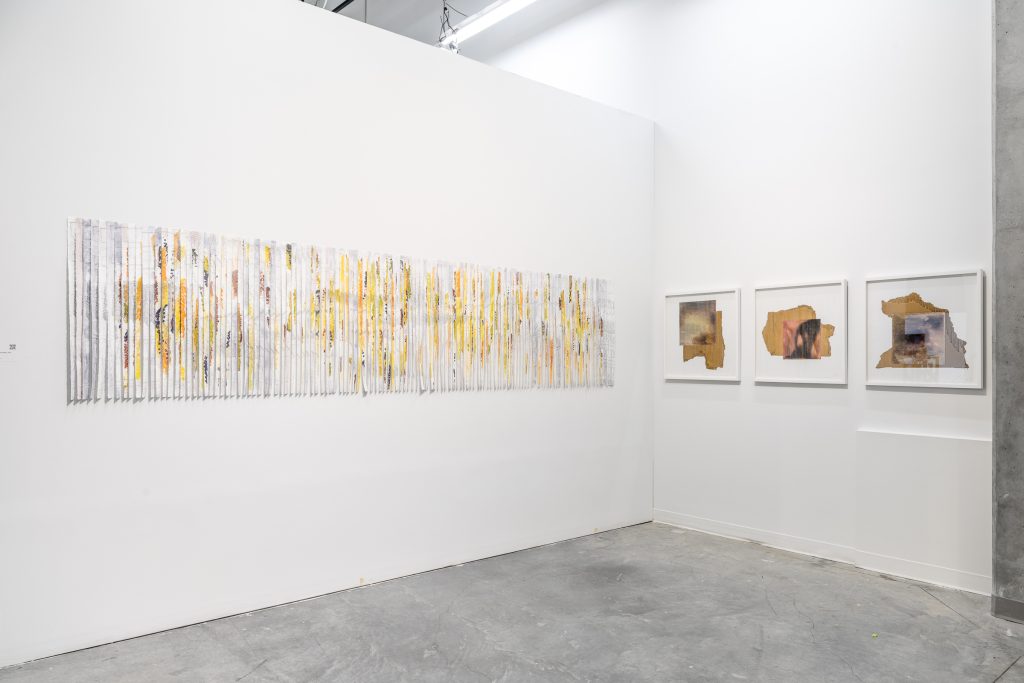 Installation view of a corner space of two walls. On the first wall is a mounted piece madd up of many vertical strips of canvas with abstract hues of blue and grey, broken up by strips of yellow and red.

On the second wall are three framed mounted pieces, each showing an abstract image with square/rectangular collage of images laid over unevenly bordered shapes. 