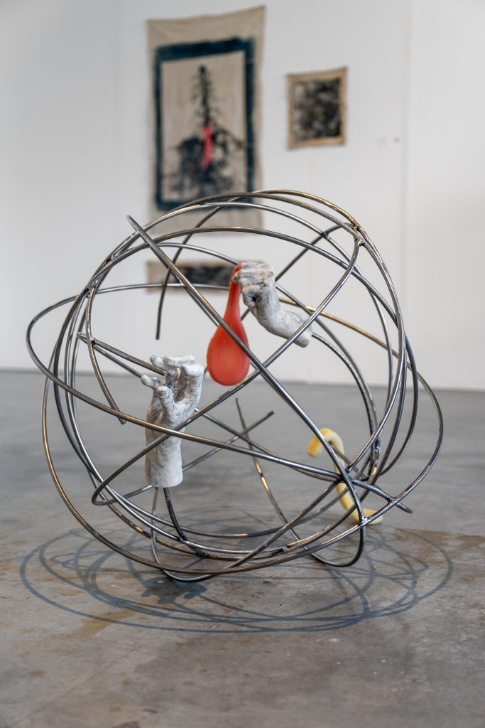 Installation view of a single physical piece on the floor, a wireframe globe with two sculptures of disembodied hands in space, one hand holds a deflated red balloon. 