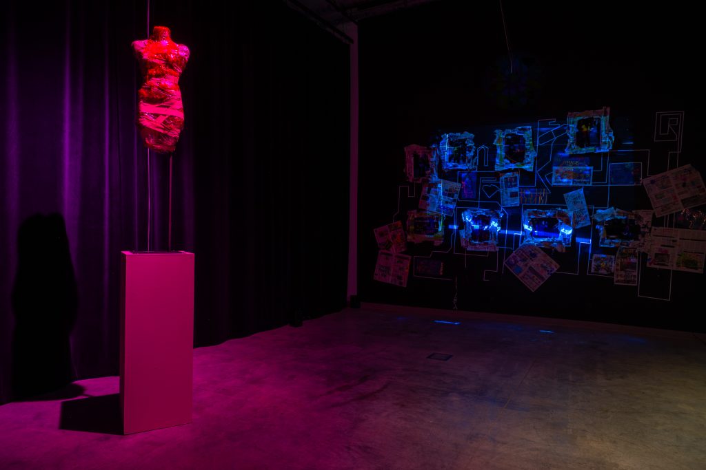 An installation involves a very dark space and light. The entire space was shrouded in black cloth, it was dim inside. On the left display was a modeling hanger with what looked like a short dress with ruffles on it, with a bandage and plush-like material. The entire modeling rack was shrouded in a pinkish-purple light. On the right side was a wall of collage, similar to a detective's clue organizer board, with pictures taped and lines drawn on it. Blue light illuminates the right side.