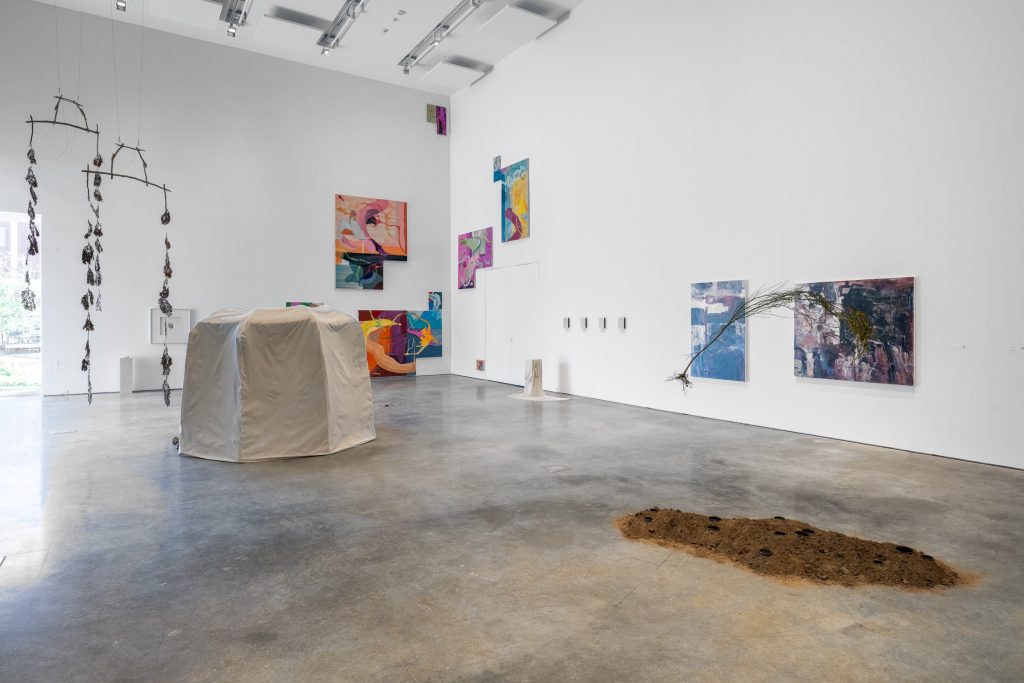 Gallery installation view. In the foreground are two hanging installations. The right side looks like the shape of a slender plant with dark brown earth and black stones beneath it. The one on the left has feathery plants hanging down from branches. Behind it is a large white installation shaped like a Cannelés. On the right, a small white column is placed on the ground and the wall next to it displays four white squares. Two paintings are also on the wall, and a group of oil paintings in highly saturated colors are placed in the back corner.