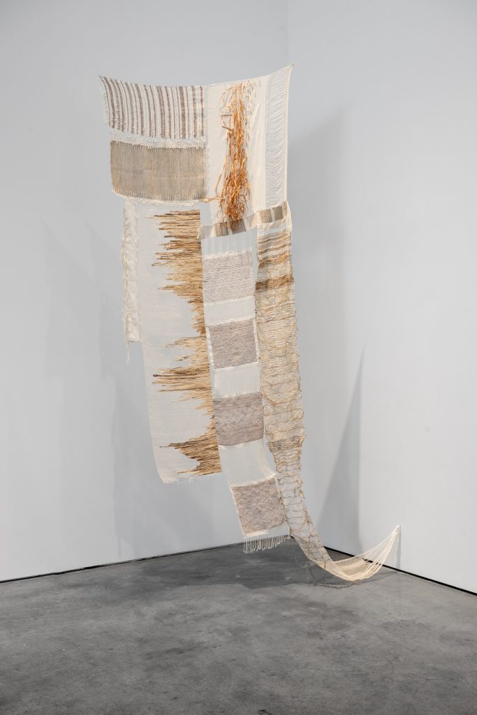 A large fabric installation. The piece is hung up with the bottom corners nailed to the side wall for better display. Light sheer gauze has light curry squares and lines stitched in the center. The left side of the gauze has lines of different lengths from top to bottom, the right side has a straw-like material on the top, while below it is made of material that mimics a translucent texture similar to layers of old book pages.