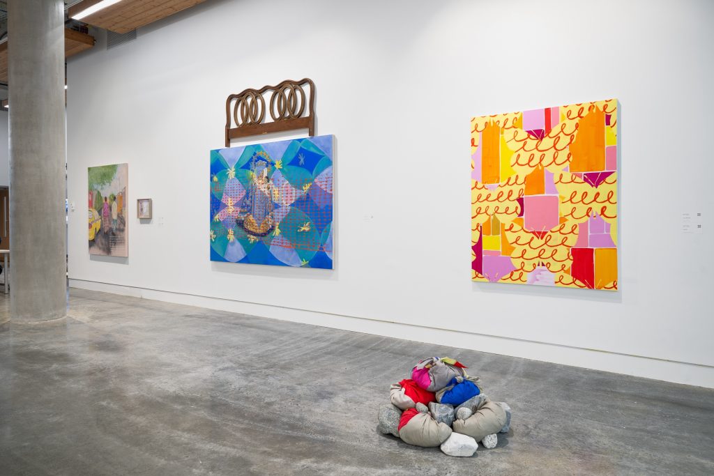 Gallery installation view. Four pieces are shown. Three pieces are paint on canvas artworks, and the fourth is an assembly of bundled red, blue, and grey fabrics on the floor. One canvas piece shows orange, red, purple, and pink geometric shapes on top of red cursive swirls, on a yellow background. The second canvas piece shows turquoise and blue overlapping shapes with bursts of yellow filaments with a grid of red points and lines overlaid. On top of the canvas is a mounted iron frame with interleaving ovals.
The third canvas is a painted scene of two people walking down the sidewalk, viewed from behind, while a yellow car passes to their left. 