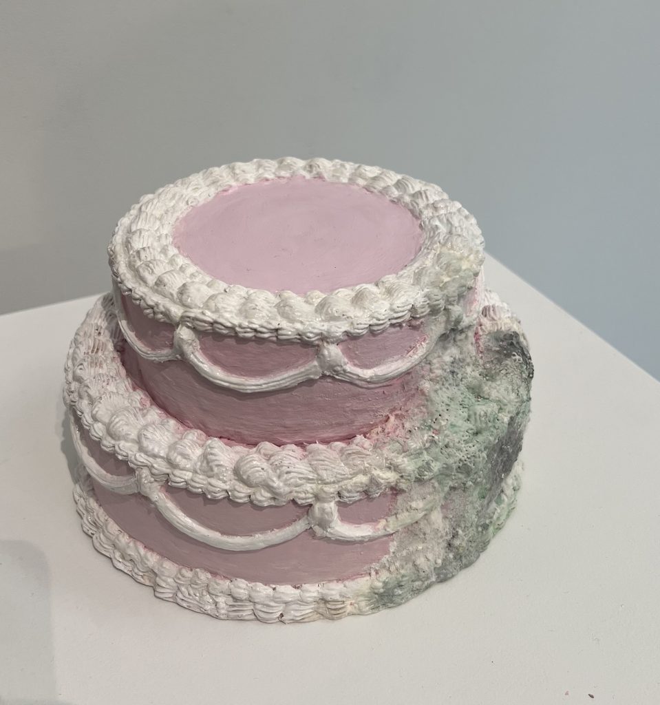 Front image of "spoiled", a pink ceramic cake decorated with detailed icing, but in the process of molding.