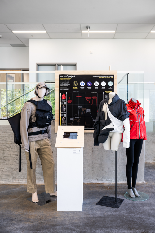 Installation view of various clothing pieces on mannequins, with a display board showing aspects of the design in the background 