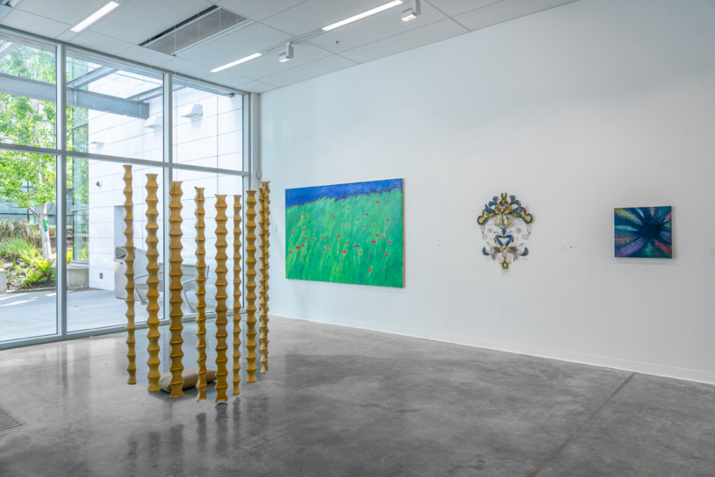 Installation view of four works.
The first work is a sculpture made of large hanging vertical strips shaped into a ripple format. 

The second piece is a wall-mounted canvas painting of a grassy field with red flowers and a strip of blue sky at the top.

The third piece is a wall-mounted sculpture of assembled glass pieces into an ornate pattern similar to a coat of arms. 

The fourth piece is a wall-mounted canvas painting depicting a number of coloured feathers arranged in a circle with their tops pointing inwards.