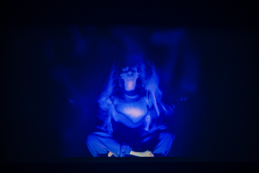 Closeup of a projected image showing a darkened space with a person sitting cross-legged and eyes closed, dressed in blue, with flares of fabric drifting upwards. 