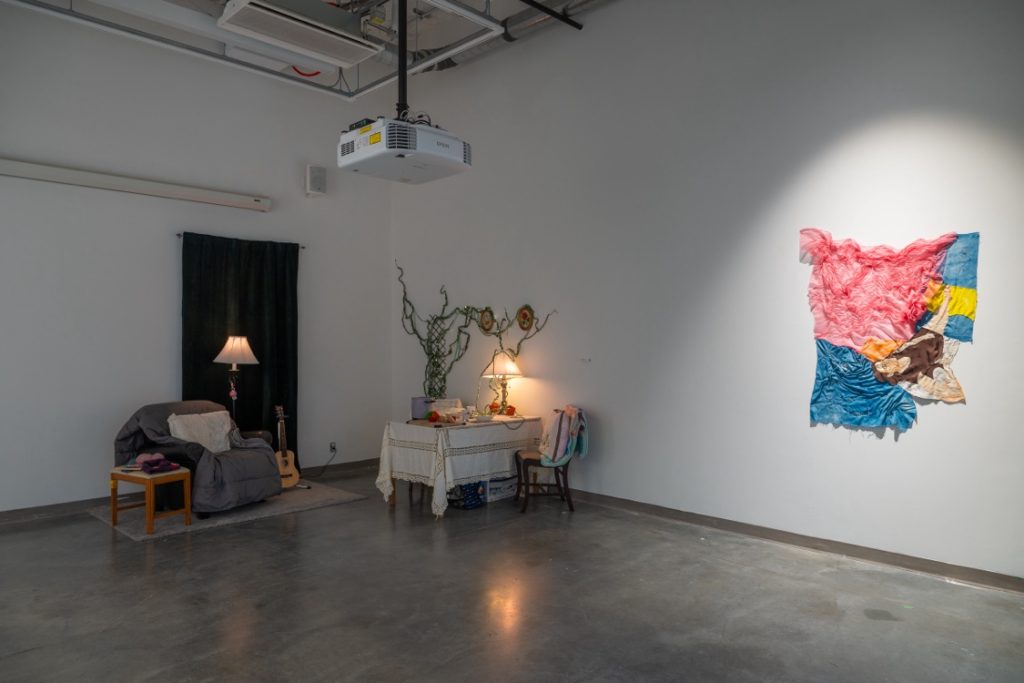 Installation view of a corner space. Mounted on one wall is a ruffled fabric piece of pink, blue, yellow, and brown, illuminated by a spotlight. Nearby is a small table with tablecloth, various items and a lamp on it, and a chair beside. Across from the chair is a vine on a trellis. On the adjacent wall is an easy chair with blanket and pillow, a guitar on one side and a side table on the other. Behind the chair is a lamp and black curtain hung on the wall. 