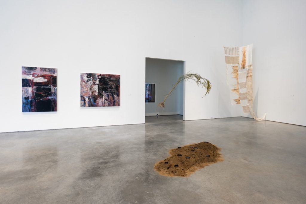 Installation view of various pieces. In the centre is a mound of dirt on the floor, with an uprooted small tree suspended above the dirt. 

In the corner is a long suspended fabric piece of white, beige, and gold. 

On the adjacent wall are two abstract painted pieces on canvas.
