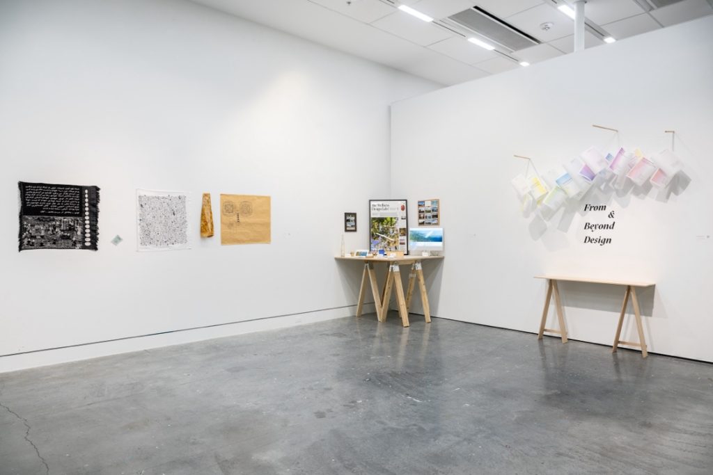 Installation view of various pieces. In the corner is a small table with various items on it. A second table is placed against a wall, with the words "From & Beyond Design" written on the wall. A collection of white and faintly coloured pieces is suspended above the writing and the table.

On the adjacent wall are three printed pieces.