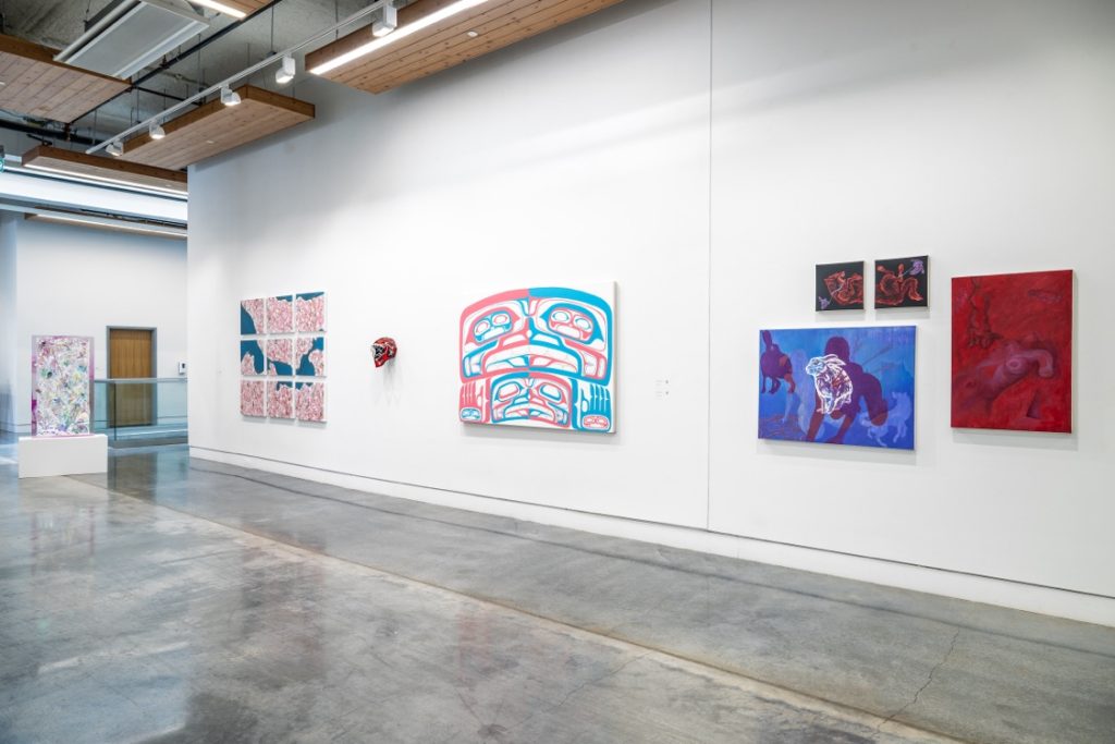 Installation view of various pieces, including a work made up of a grid of 9 small square canvasses, a Coast Salish style painting in red and blue, a canvas with the drawing of a white rabbit against a silhouette of a person on hands and knees, on a blue background. Beside it is a canvas with a torso painted in red, and two smaller canvases that form one larger image.