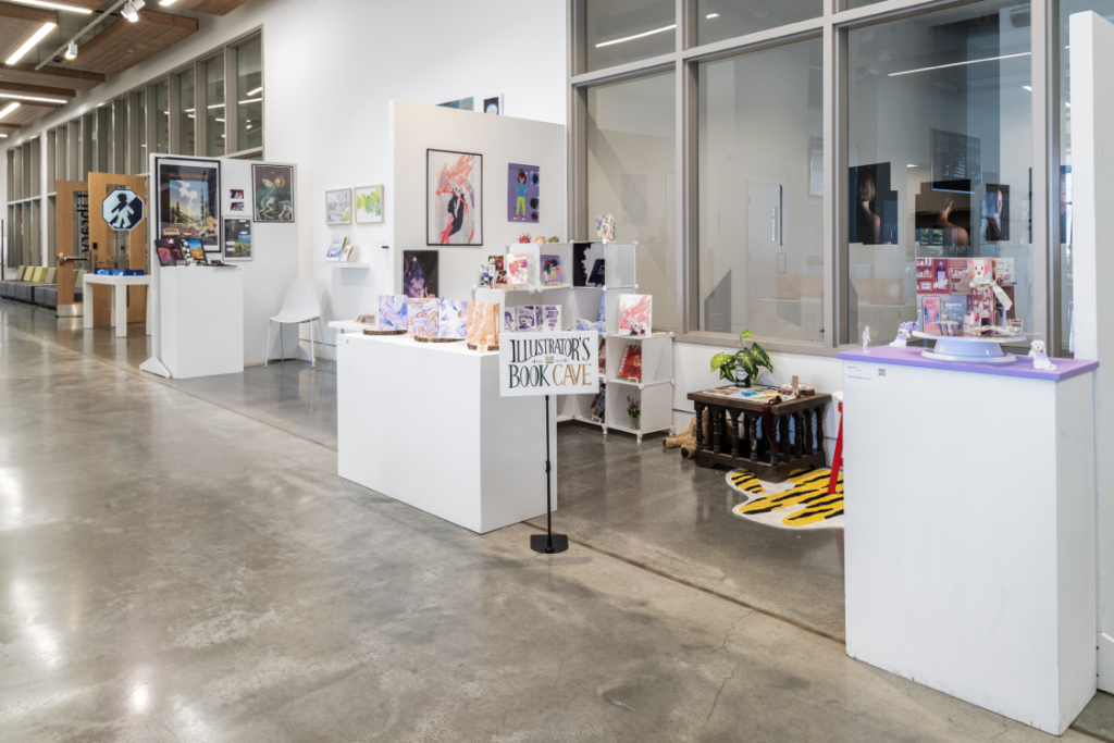Installation view of several projects, in the foreground is a space with a sign reading "Illustrator's Book Cave" and holding an assortment of artifacts and books on stands.