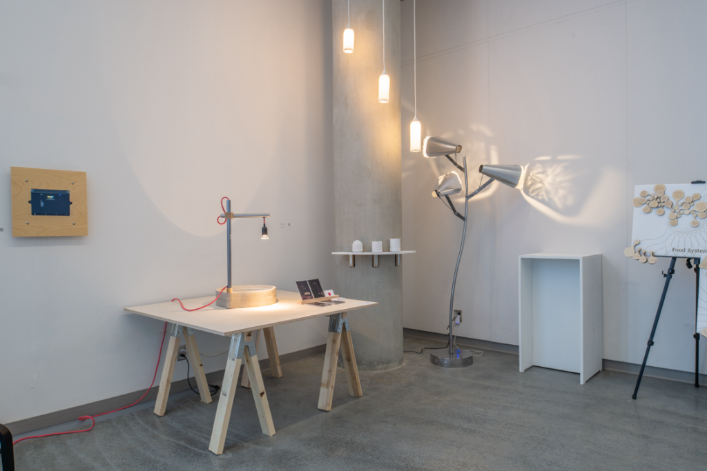 Installation view of various light fixtures with pieces arranged on a table, shelf, and tripod. One light fixture projects textured shadows on the wall.