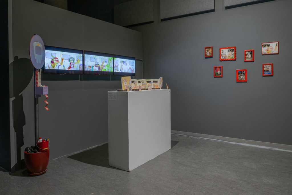 Installation view in a darkened corner space. Three video displays are mounted on the wall showing parts of a long horizontal scene. A free standing pole with a bus icon on top has a ticket dispenser attached to it. A podium in front of the displays holds am illustrated cardboard display. On the second wall are a number of small pictures in bright red frames