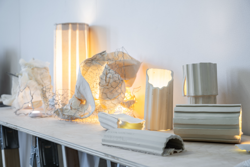 A collection of white sculpture pieces arranged on a table. Several pieces are shaped as hollow classical pillars, some with lights shining from within. A centrepiece of twisted wire framing around folded paper, lit from within.