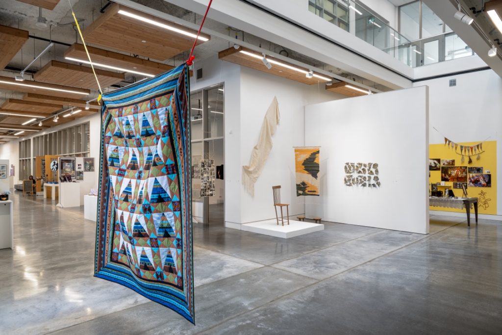 Installation view of several pieces, with a quilted blanket suspended in the foreground