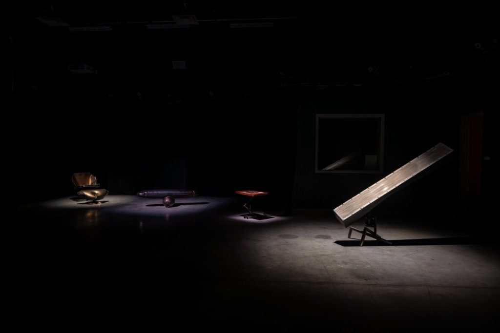 Installation view of a darkened room with four pieces illuminated by spotlights. One piece is a large wooden slab on an angle, and three others of abstract form.