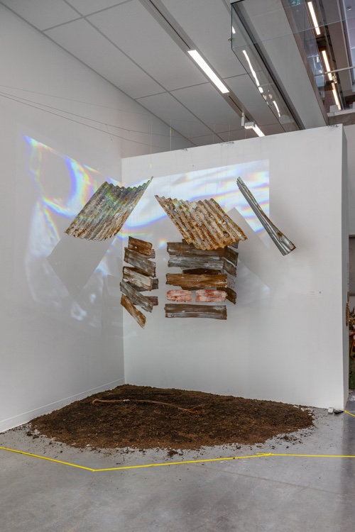 Installation view in a corner space demarcated with a yellow line on the ground. Within the yellow line is some dirt, and suspended above the dirt are pieces of sheet metal and wood. Illumination is reflected in iridescent tones on the wall behind. 
